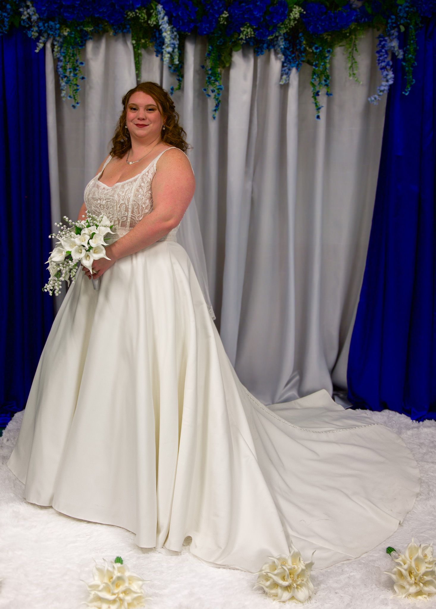 Bride in elegant white satin ball gown with beaded bodice holds bouquet before blue floral backdrop at wedding.