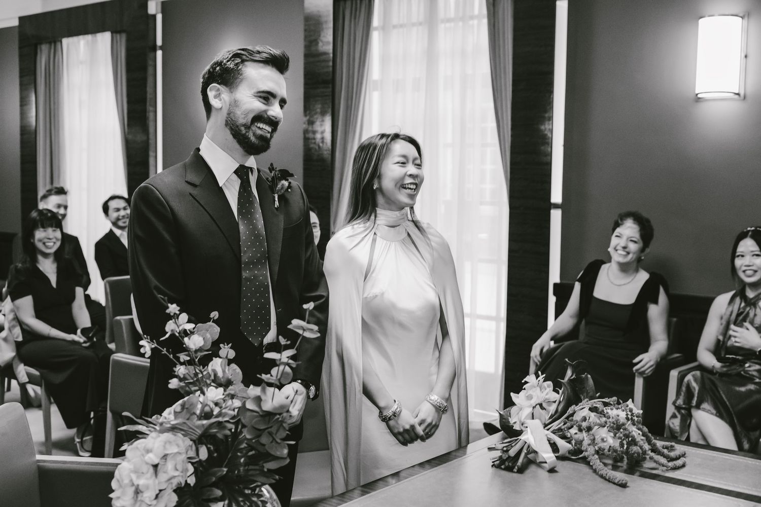 Black and white wedding ceremony photo of smiling couple standing at altar with guests seated in background.