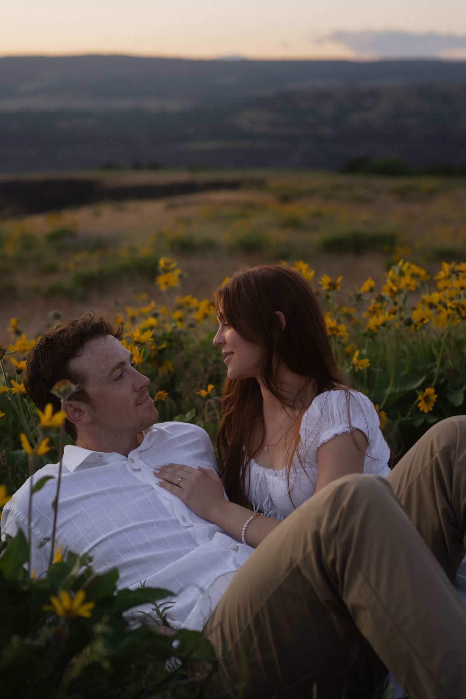 A couple sits together in a field of yellow wildflowers at sunset, gazing at each other romantically.