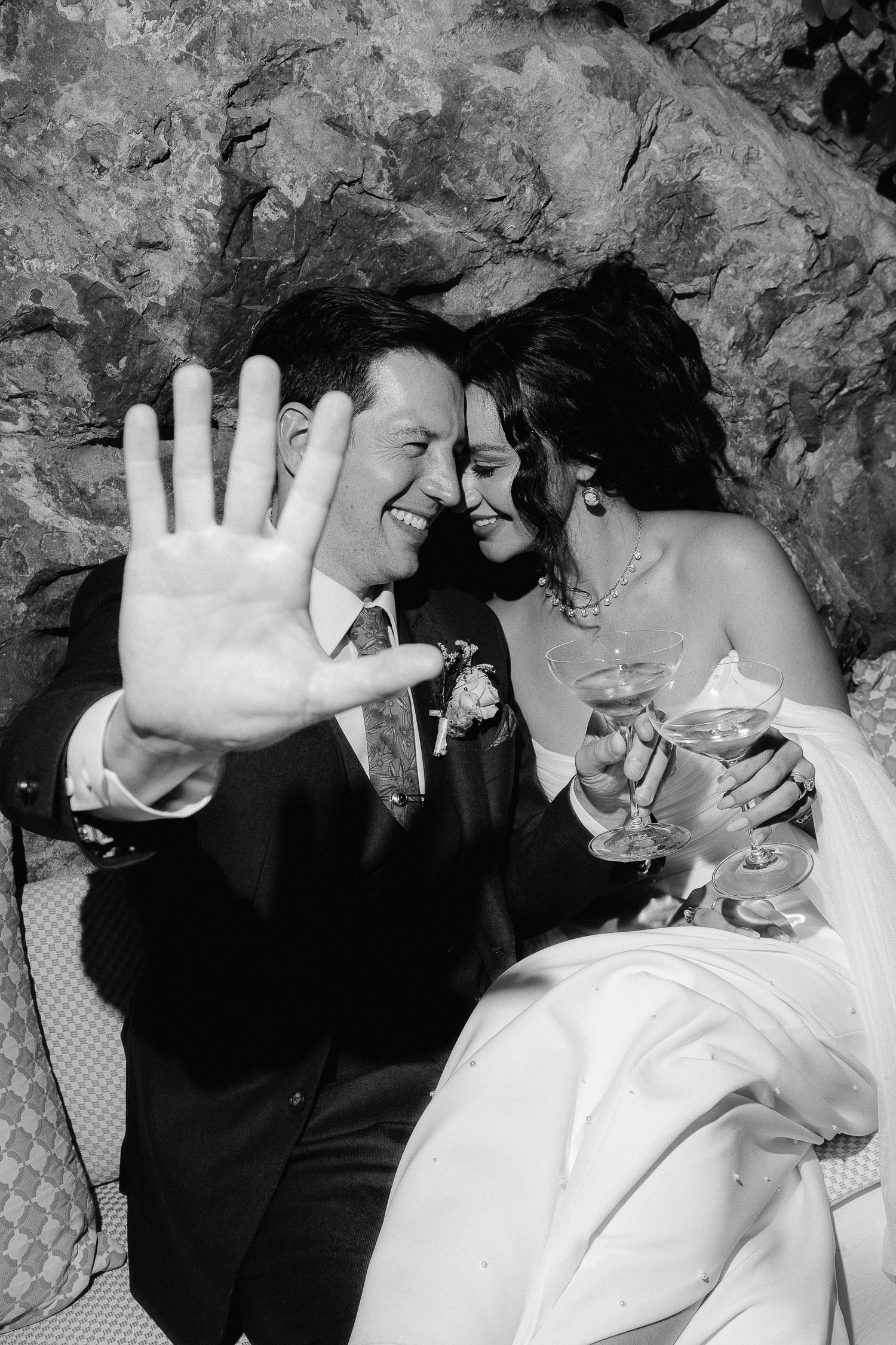 Black and white photo of a laughing groom waving at camera beside his smiling bride holding champagne glasses.