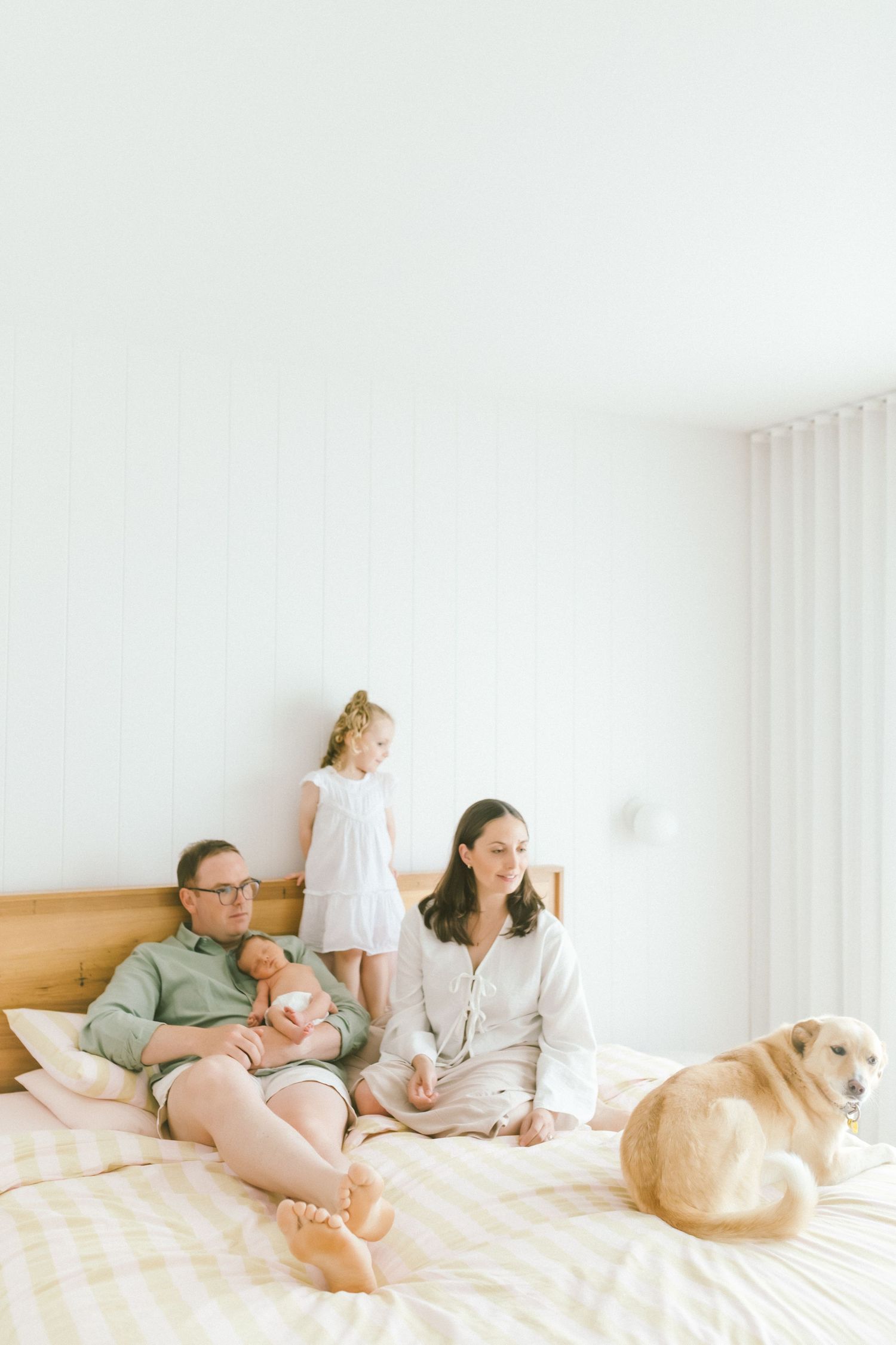 Family with newborn, toddler, and golden dog relaxing on a bright white bedroom bed in soft natural light.