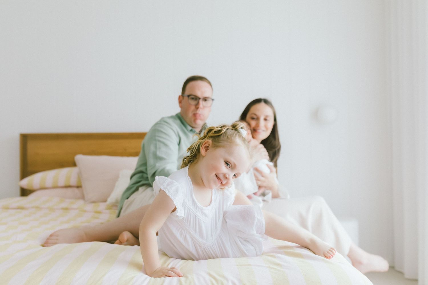 Family of three relaxing on a white bed, parents smiling behind toddler girl in white dress with floral crown.