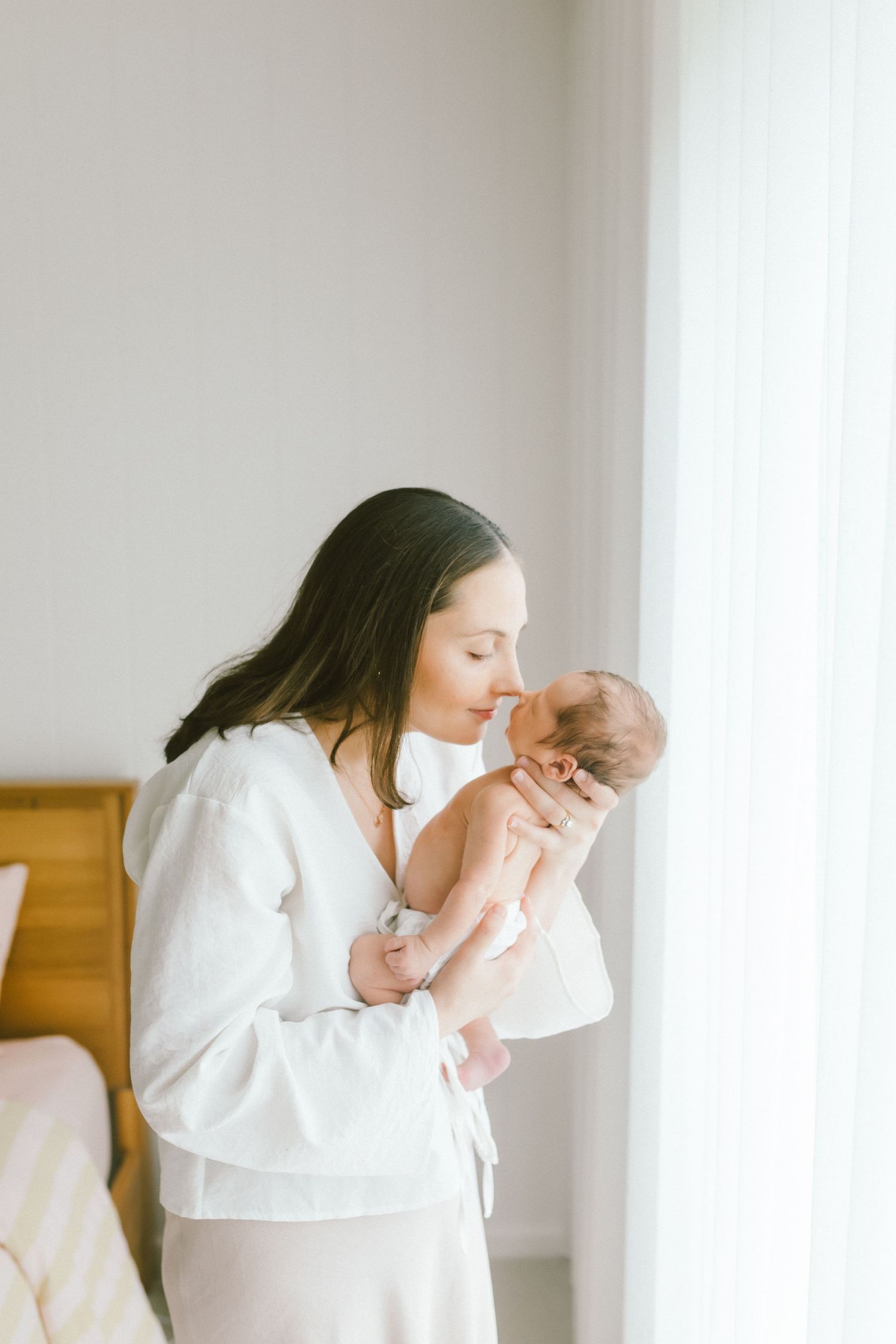 A young mother tenderly kisses her newborn baby while standing near a bright window in a softly lit room.
