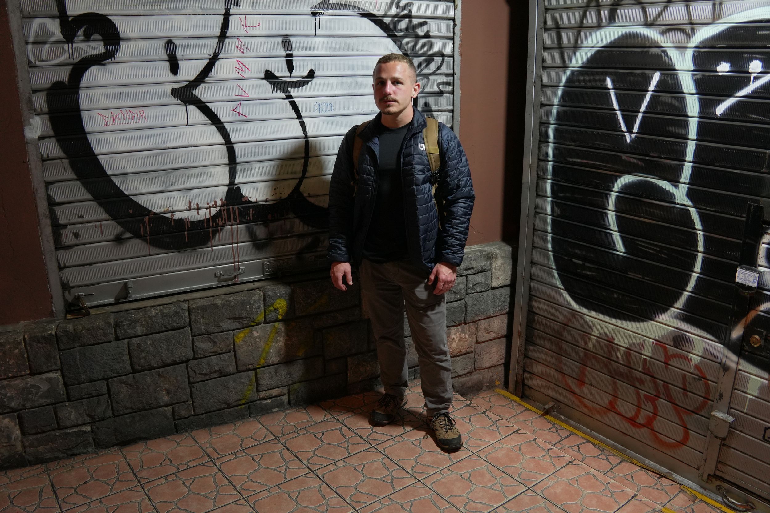 Man standing against a graffiti-covered wall in an urban setting at night.