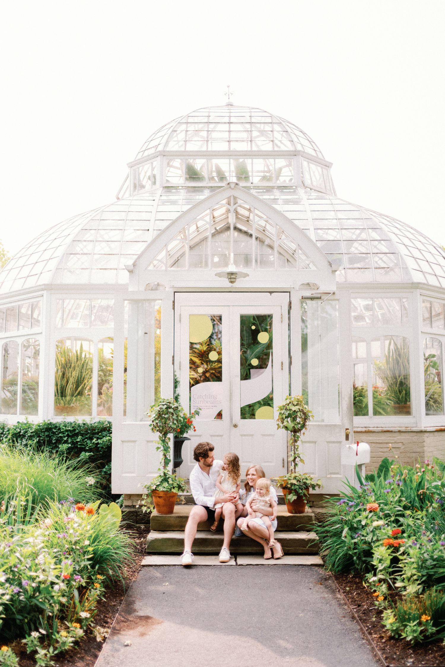 Family sitting on steps before a white Victorian greenhouse surrounded by lush gardens and colorful flowers.