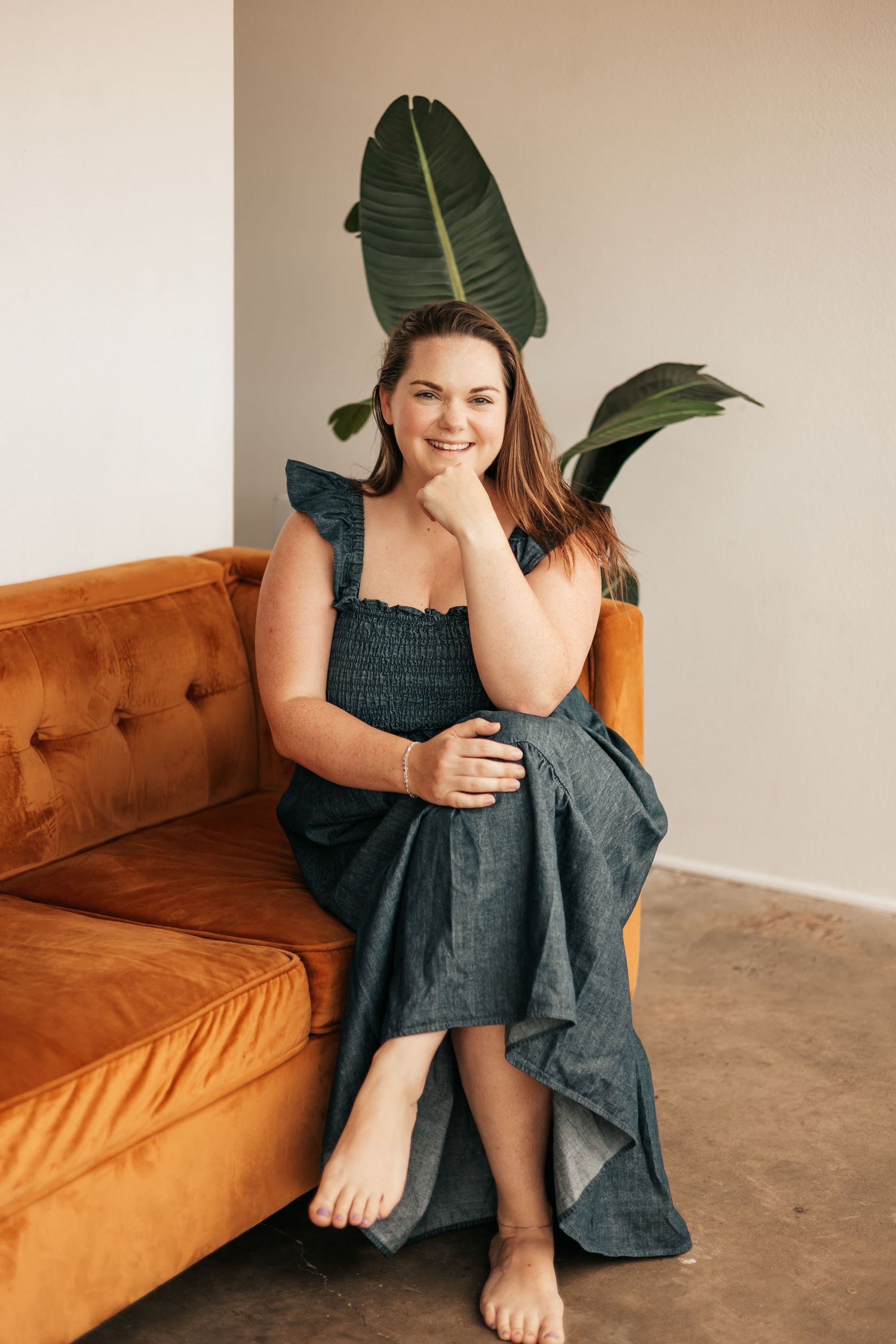 Woman in teal dress sitting barefoot on orange velvet sofa, smiling, with tropical plant behind her  | Photography Business Coach