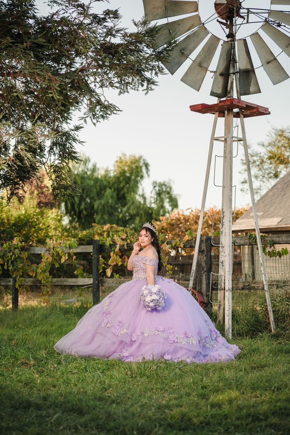 Young woman in lavender quinceañera ball gown poses near rustic windmill in lush green garden setting.