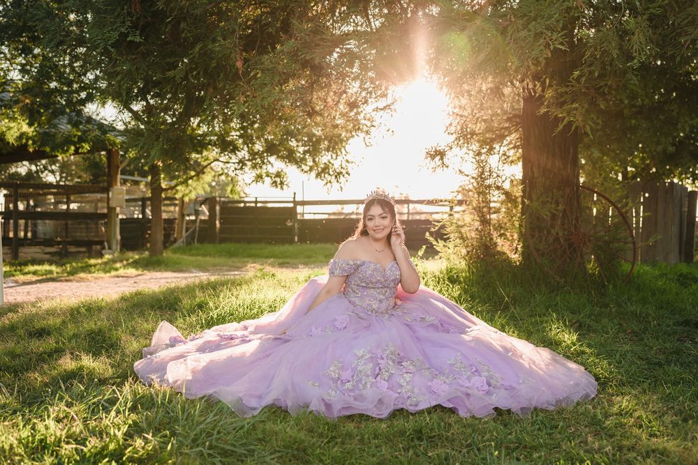 Young woman in lavender quinceañera ballgown sitting on grass in golden sunlight beneath trees.