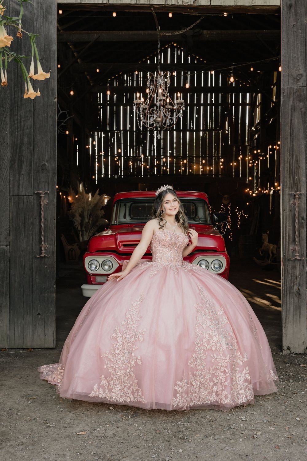 Young woman in pink ball gown quinceañera dress poses in barn doorway with red vintage truck and chandelier backdrop.