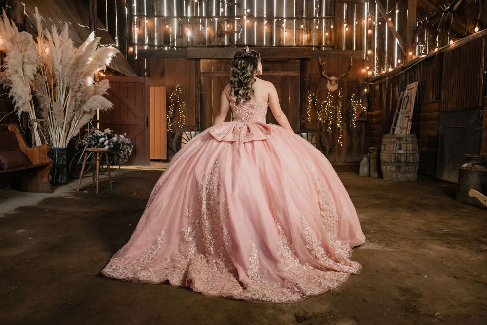 Young woman in a pink ballgown quinceañera dress poses in a rustic barn with fairy lights and floral decor.