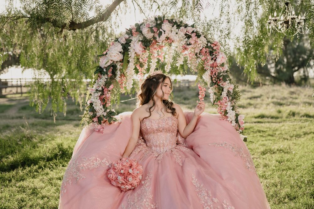 Young woman in pink quinceañera ball gown holding bouquet, seated under floral arch with willow trees.