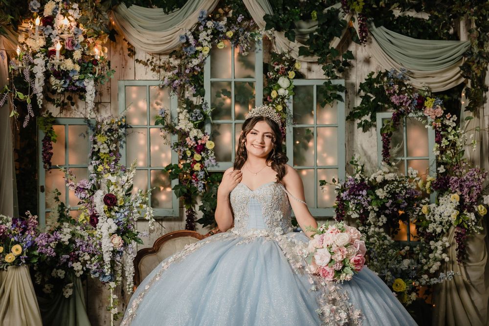 Young woman in a light blue ball gown quinceañera dress poses among lush floral arrangements in an elegant garden setting.