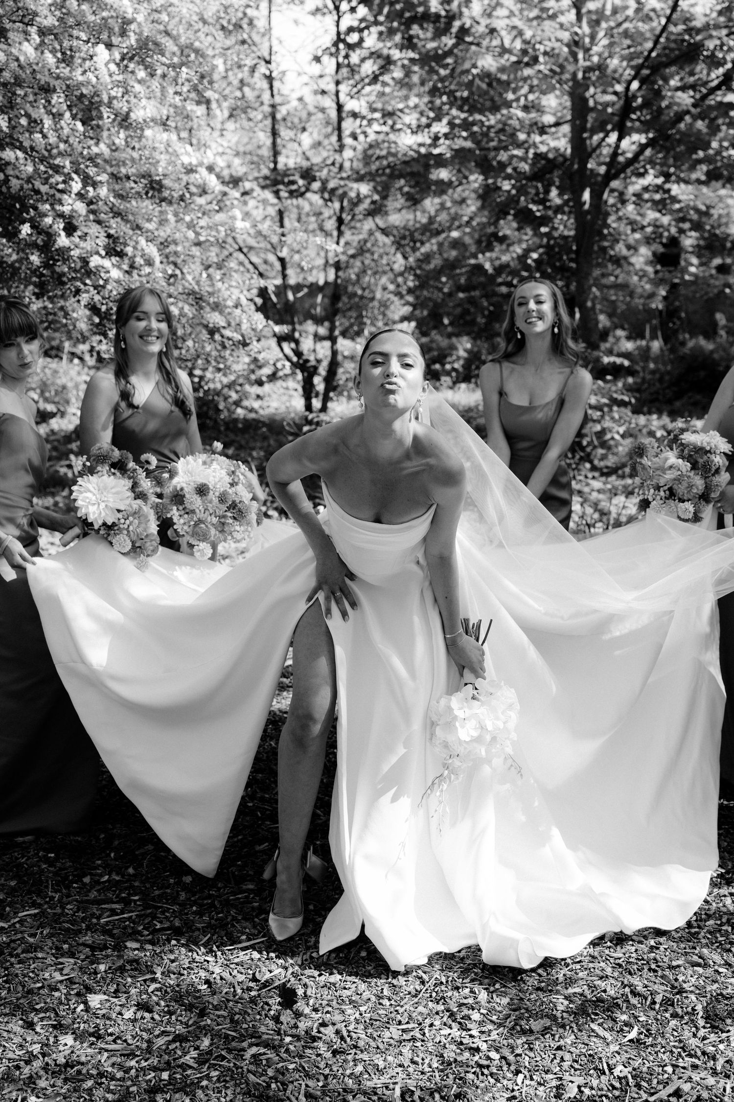 Black and white photo of a bride in a flowing gown posing with bridesmaids holding bouquets in a lush garden setting.