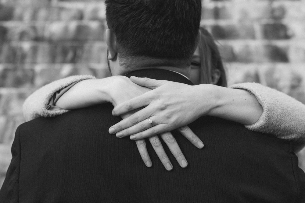 Black and white photo of a couple embracing, woman's hand with ring resting on man's shoulder.