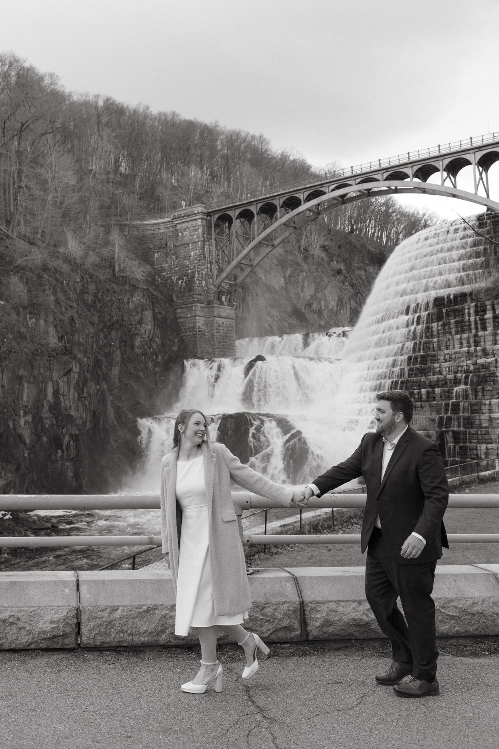 Black and white photo of a couple holding hands in front of a dramatic waterfall and arched bridge.