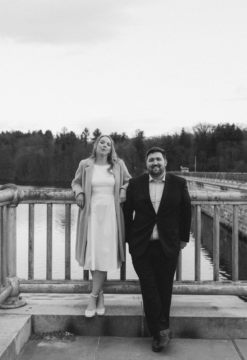 A couple poses on a bridge near a lake, woman in white dress and coat, man in dark suit, black and white photo.