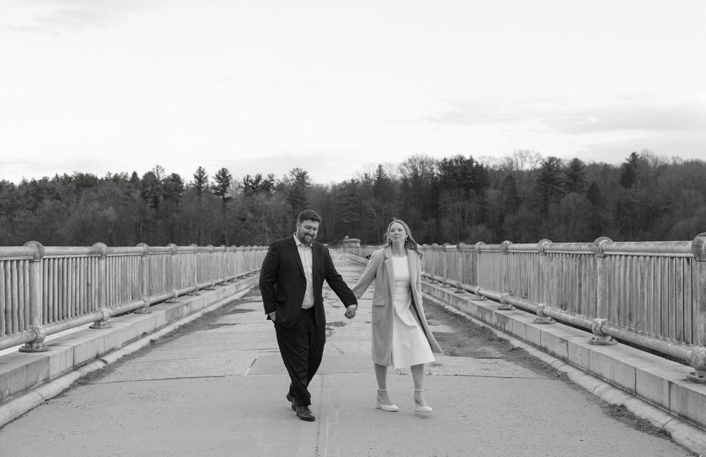 Black and white photo of a couple holding hands walking on a bridge surrounded by trees.