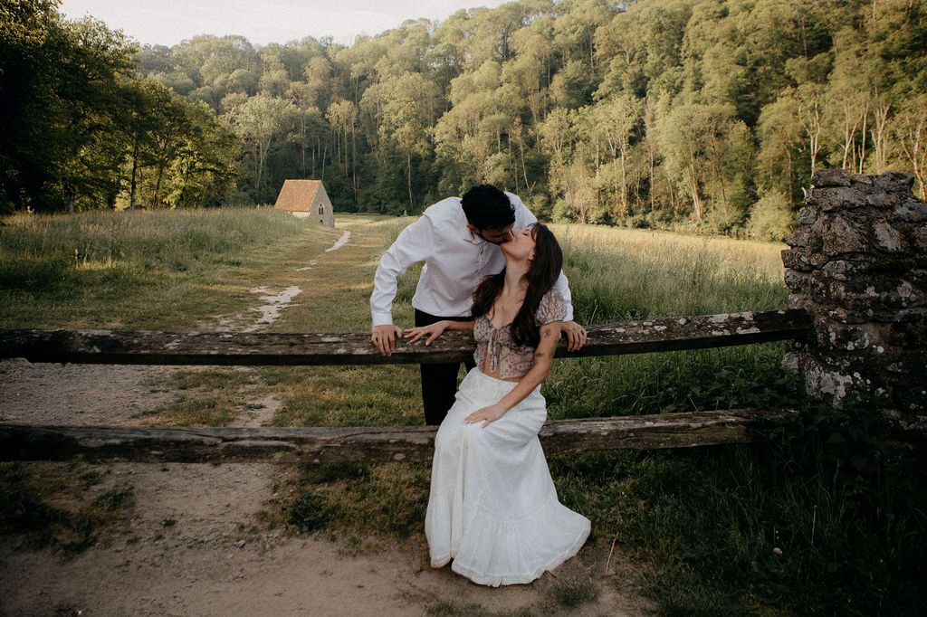 A couple shares a romantic kiss leaning over a rustic wooden fence in a scenic green countryside setting.