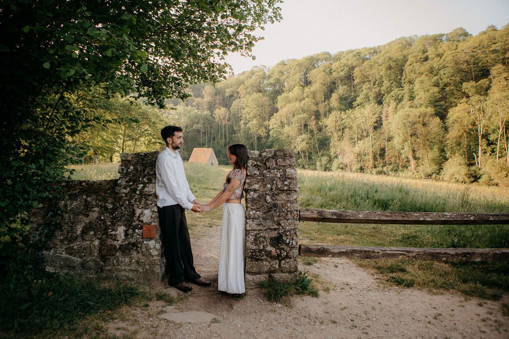 A couple holds hands while standing by an ancient stone wall surrounded by lush green forest and open fields.