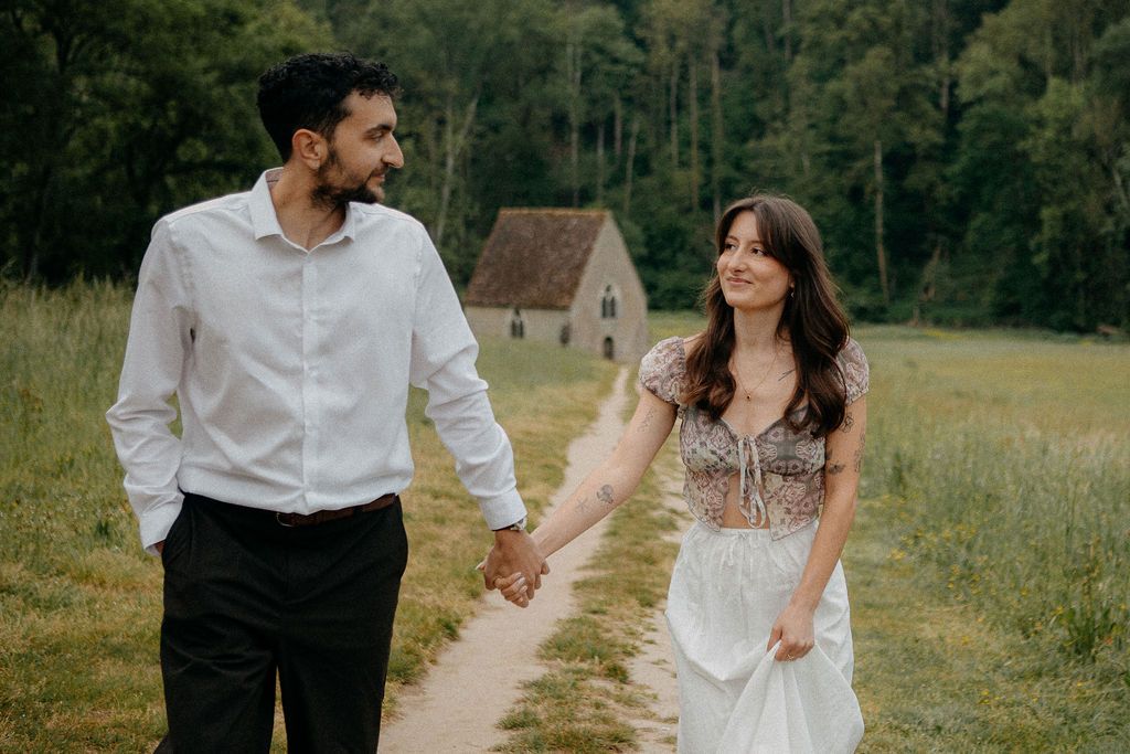A smiling couple walks hand in hand along a dirt path through a green meadow with a stone chapel behind them.