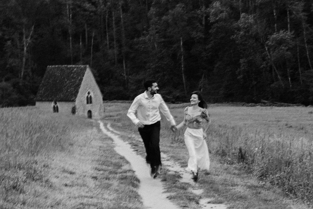A couple runs joyfully hand in hand through a meadow with a historic stone chapel in black and white photography.
