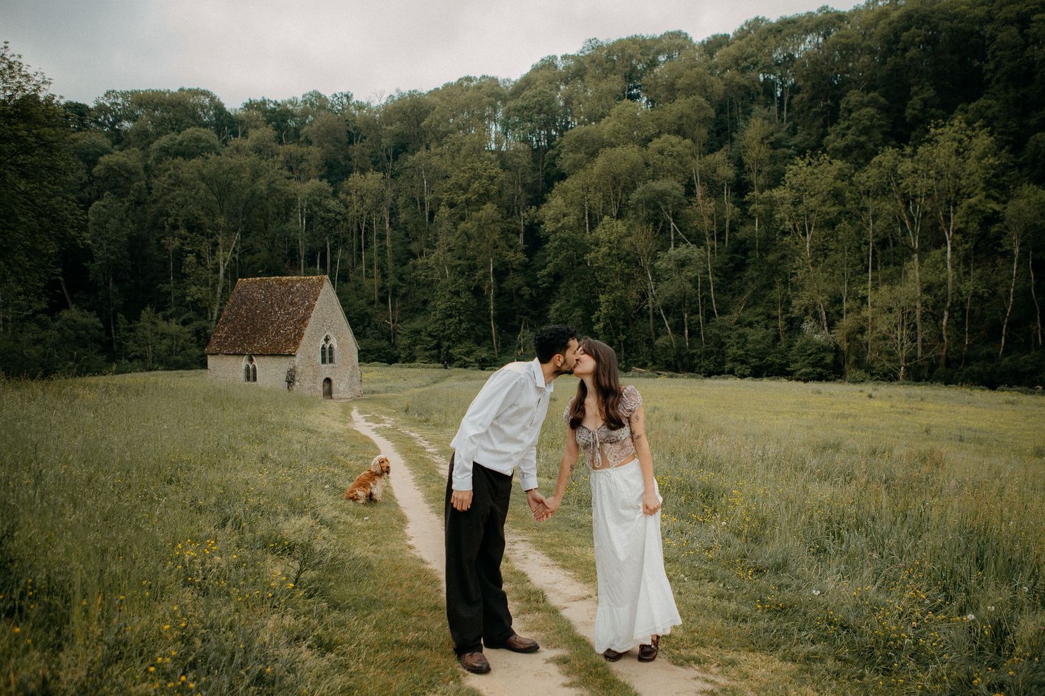 A couple shares a gentle kiss on a dirt path in a green meadow while a golden dog rests nearby.