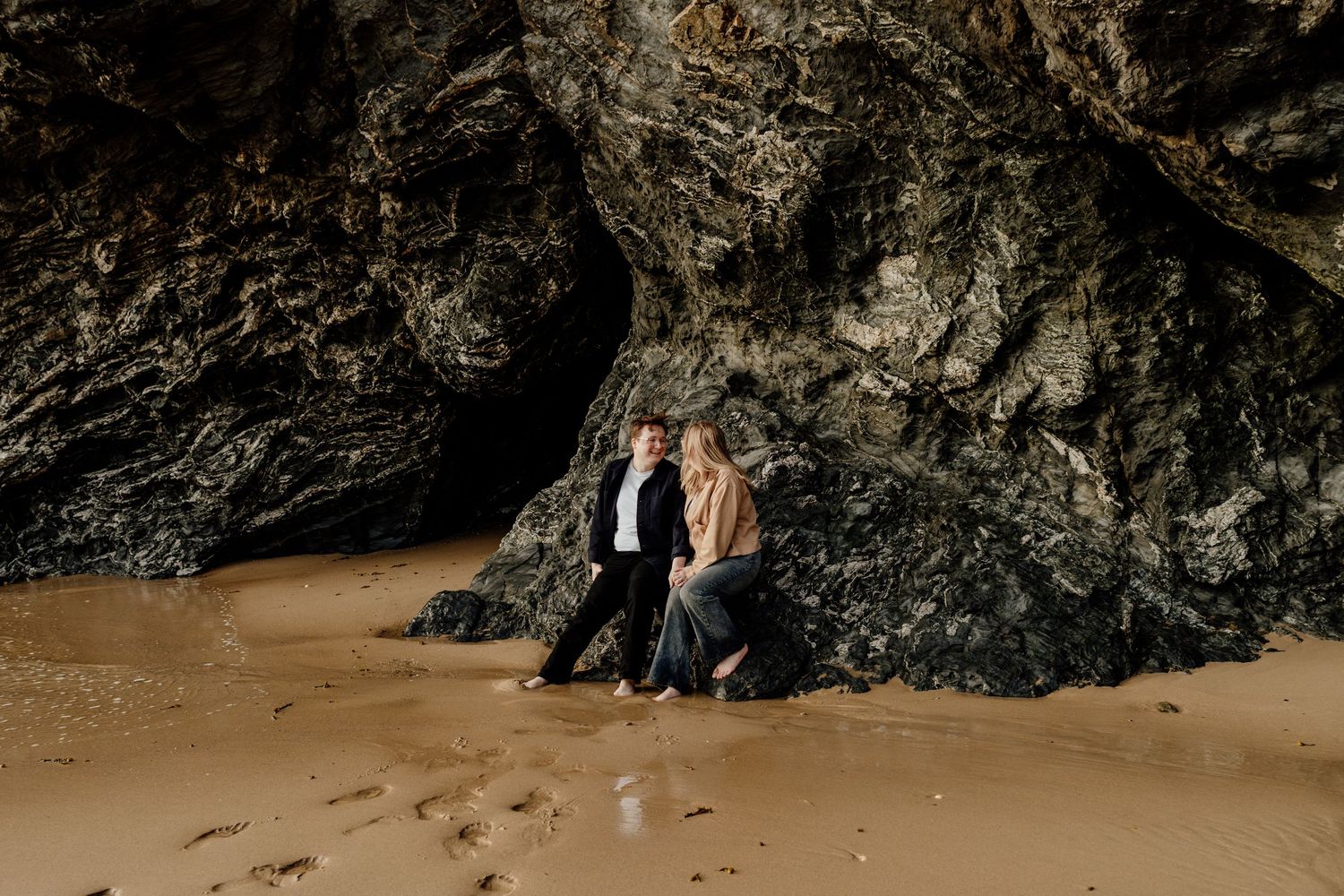 Couple leaning against rocky sea cave on sandy beach during romantic coastal engagement photo session.