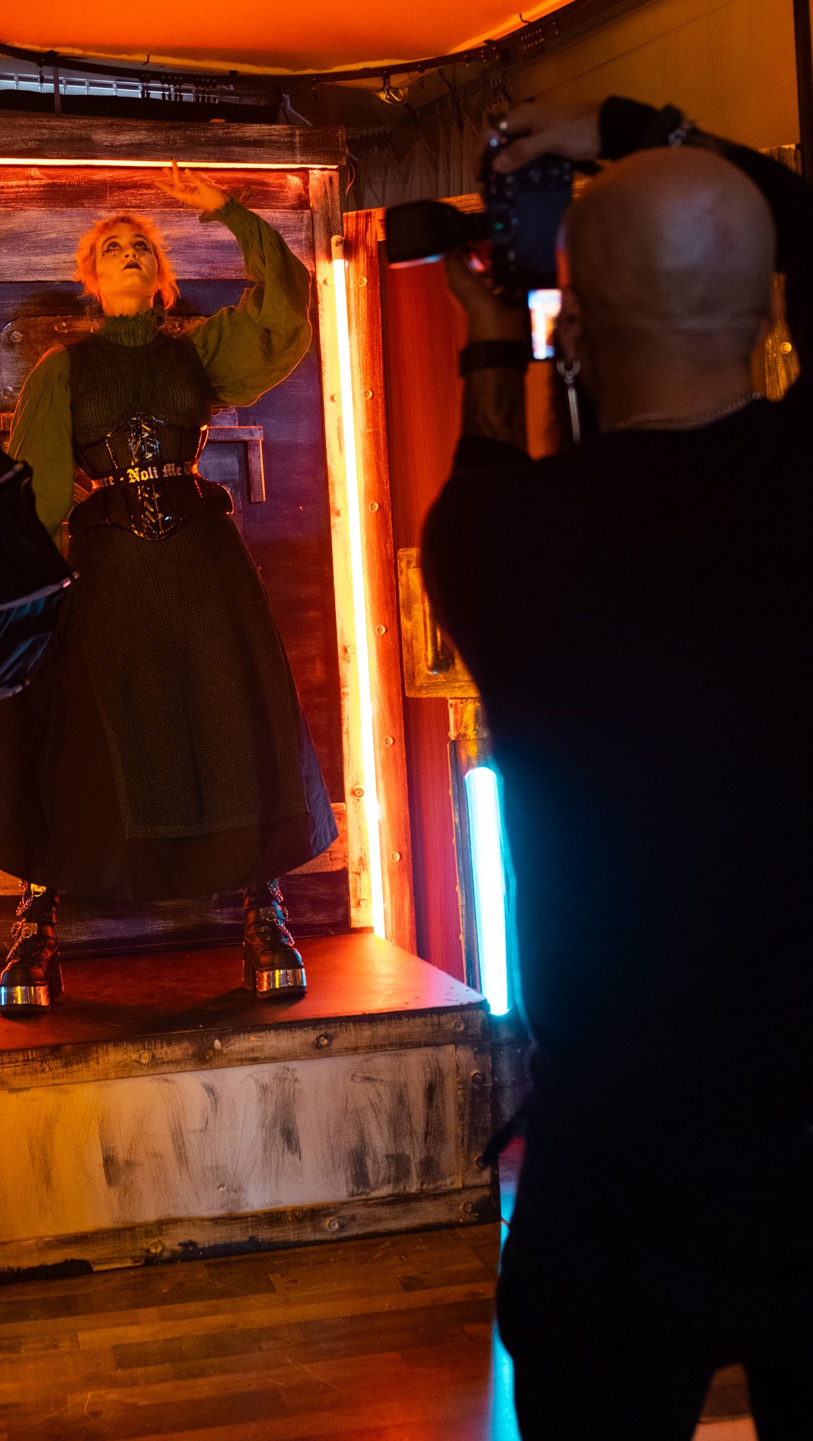 Bald photographer captures a blonde performer in green top and black skirt under neon lights on stage.