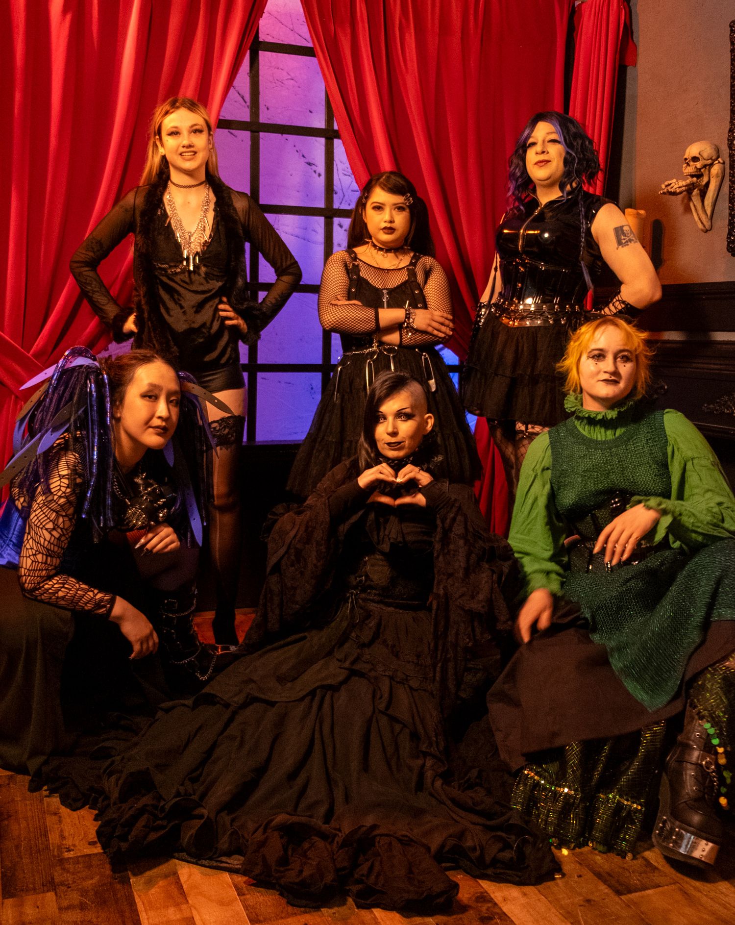 Six women in gothic black and green outfits pose dramatically in a red-draped room with moody lighting.