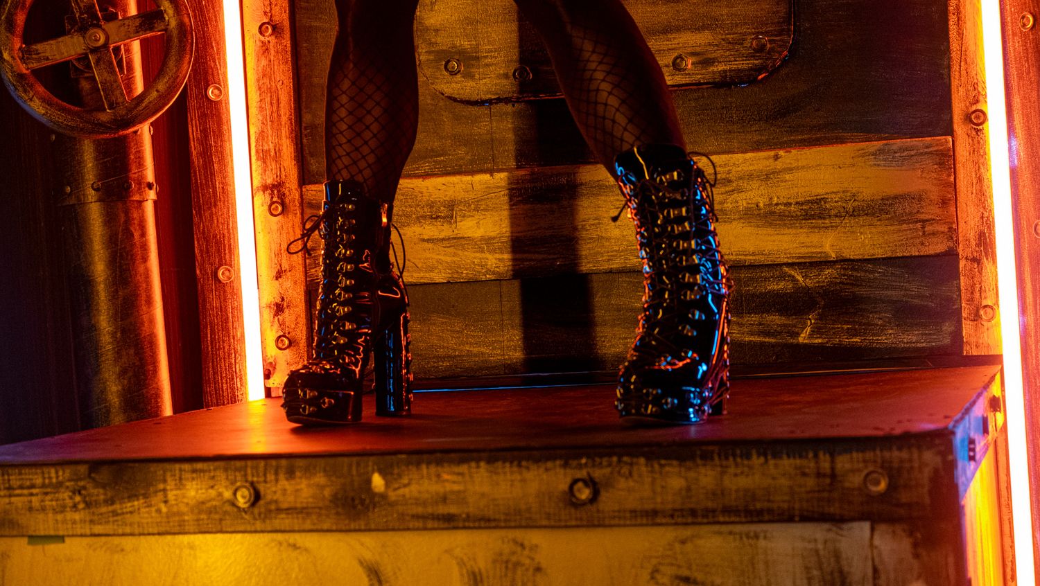 Legs in fishnet stockings and black platform lace-up boots standing on a wooden shelf with neon orange lighting.