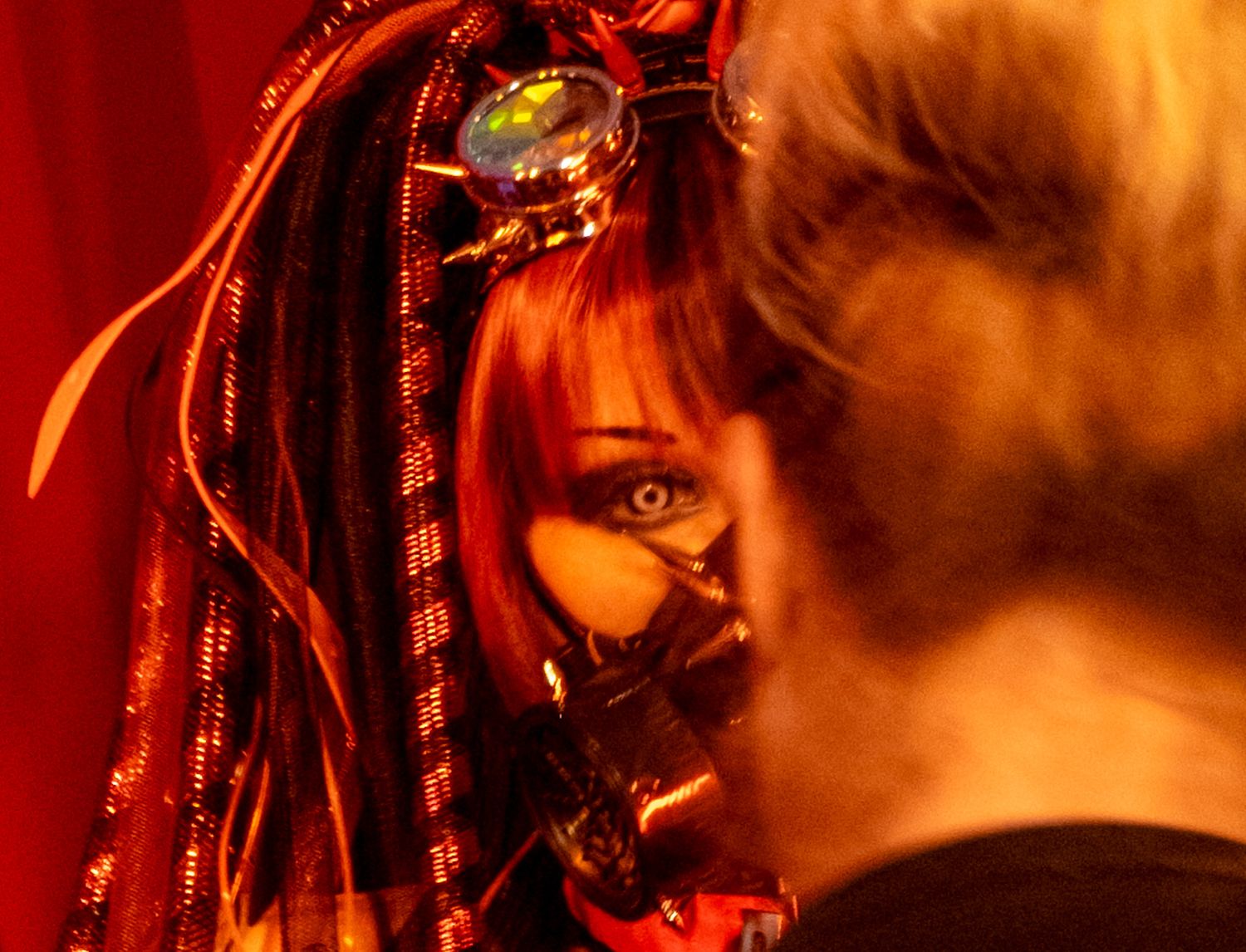 Close-up of a woman in elaborate red steampunk costume with decorative headpiece and face mask under red lighting.