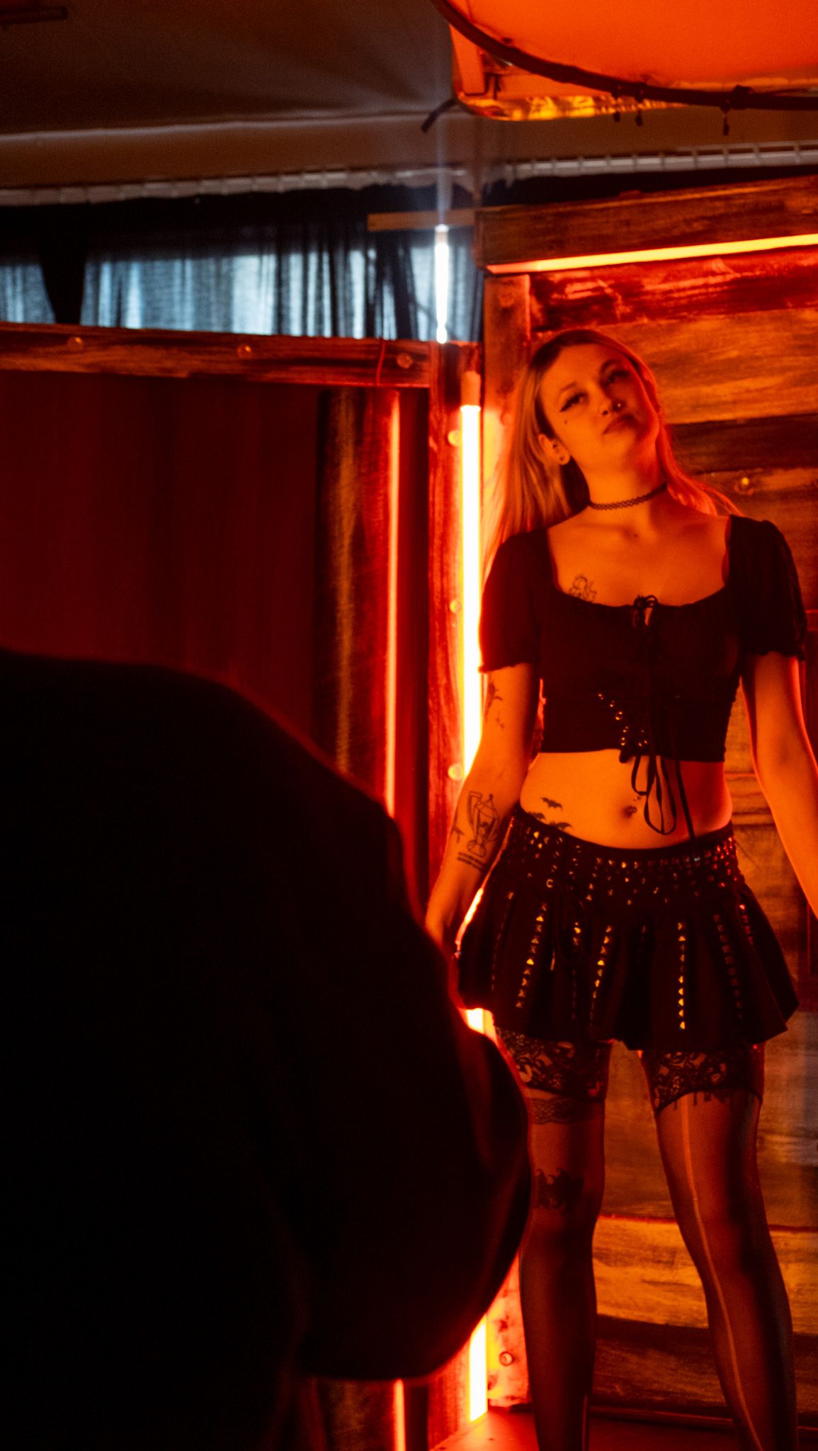 Young woman in black crop top and mini skirt poses under dramatic red neon lighting indoors.