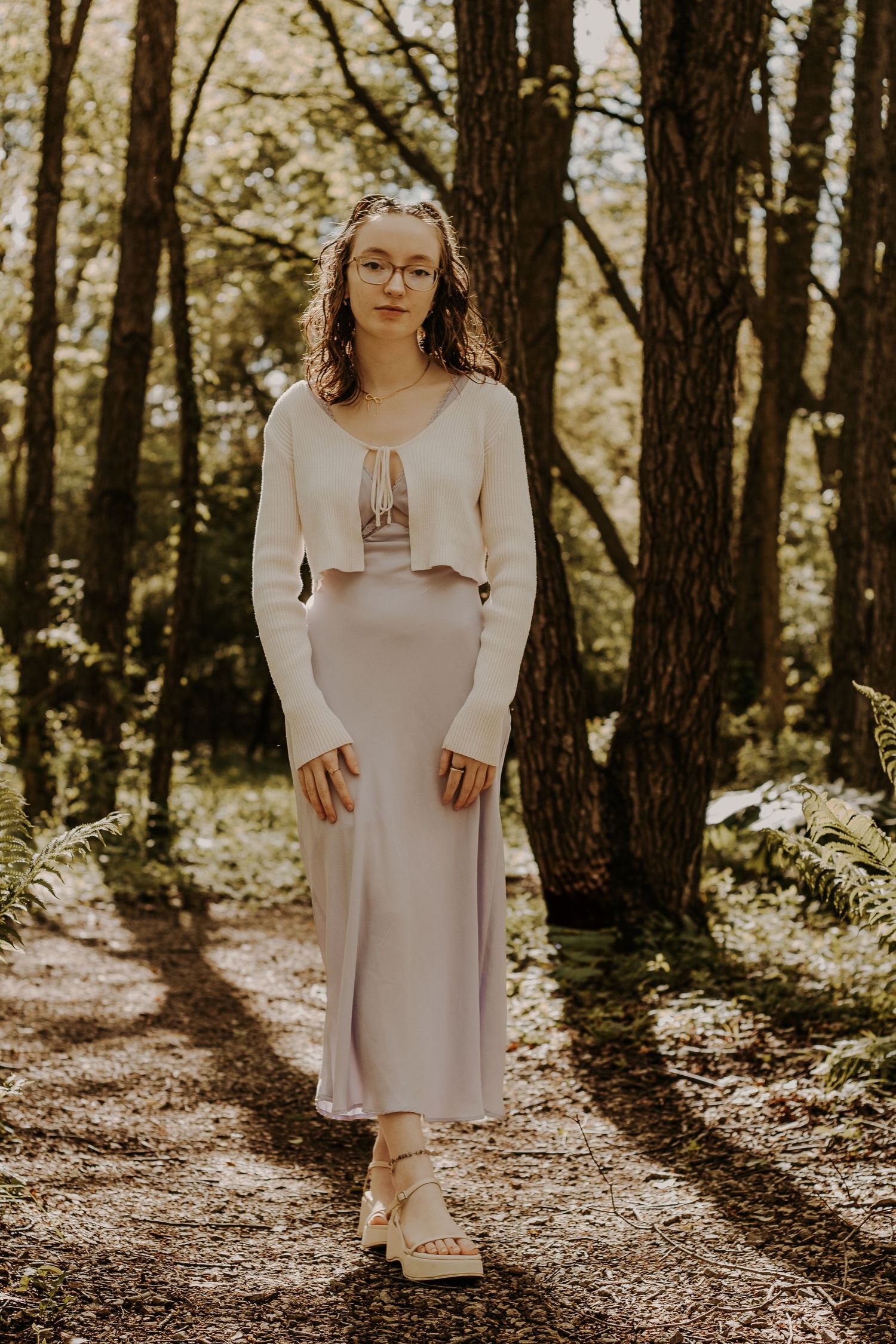 Young woman in cream cardigan and lavender midi skirt poses on a forest path surrounded by lush green trees.