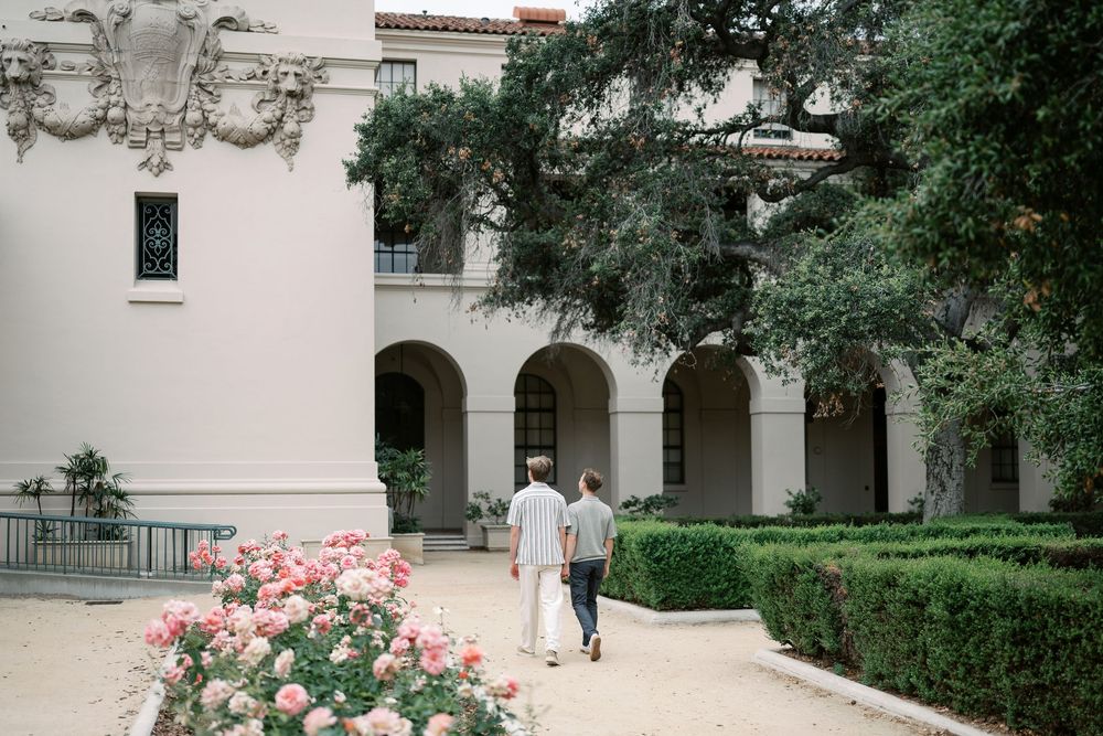 Couple walking along a garden path beside a white Spanish colonial building with arched walkways and rose bushes.