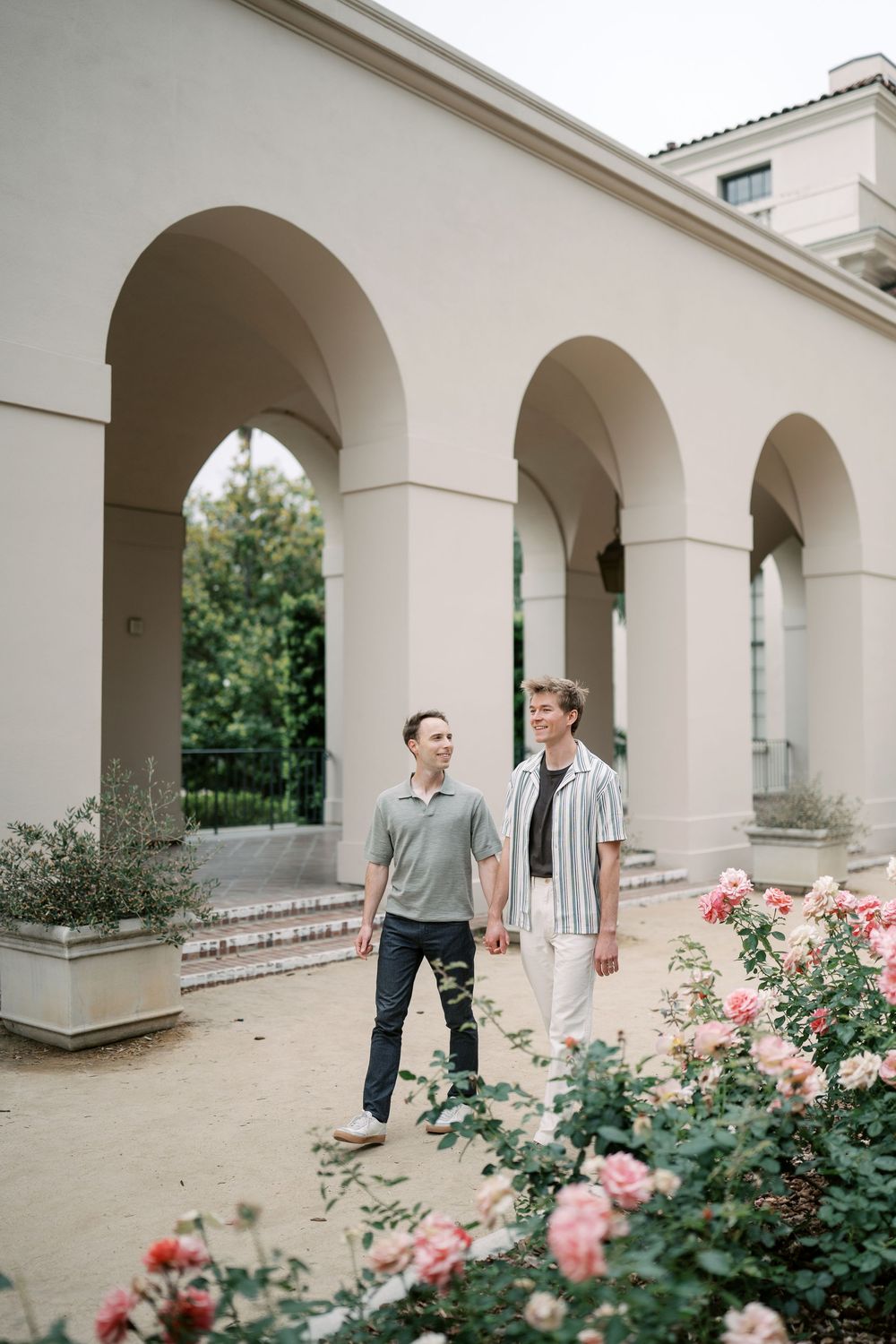 Two men hold hands walking past rose bushes beside elegant arched colonnade building.