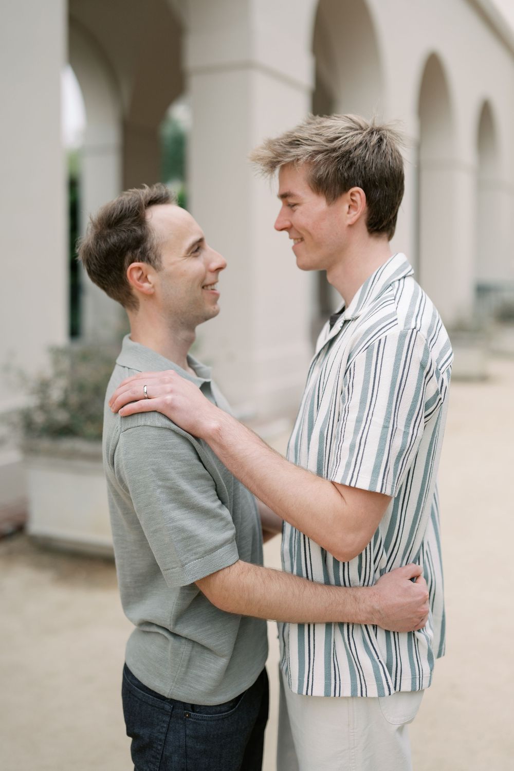 Two men embrace and smile at each other outdoors near elegant white arched columns.