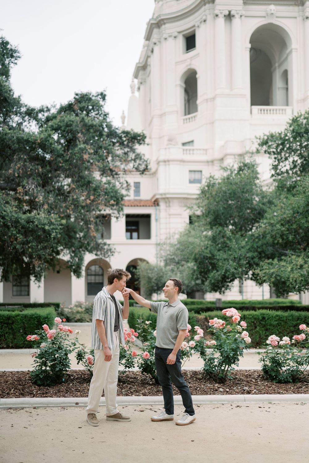 Two men share a romantic moment among pink roses outside a grand white neoclassical building.