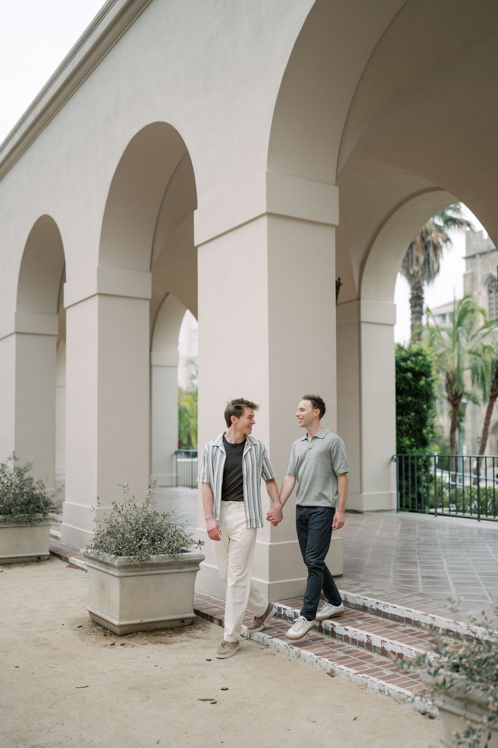 Two men hold hands and share a loving glance while walking under elegant white arches outdoors.