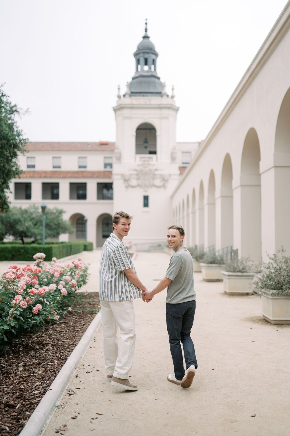 Two men hold hands walking through a courtyard with arches, roses, and a historic tower in the background.