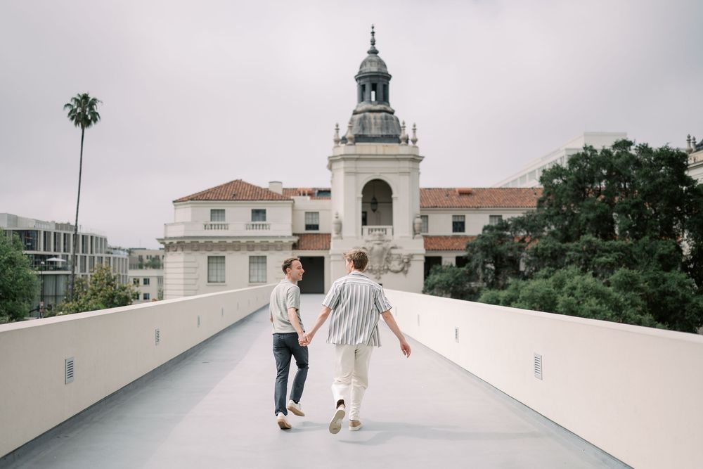 Couple holding hands walking toward historic Pasadena City Hall on a bridge, with palm trees and cloudy sky.