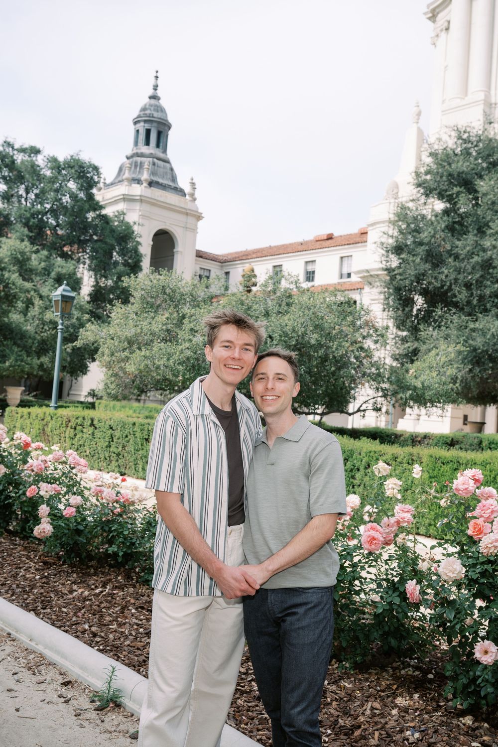 Smiling same-sex couple pose hand-in-hand among pink roses in front of a historic building.