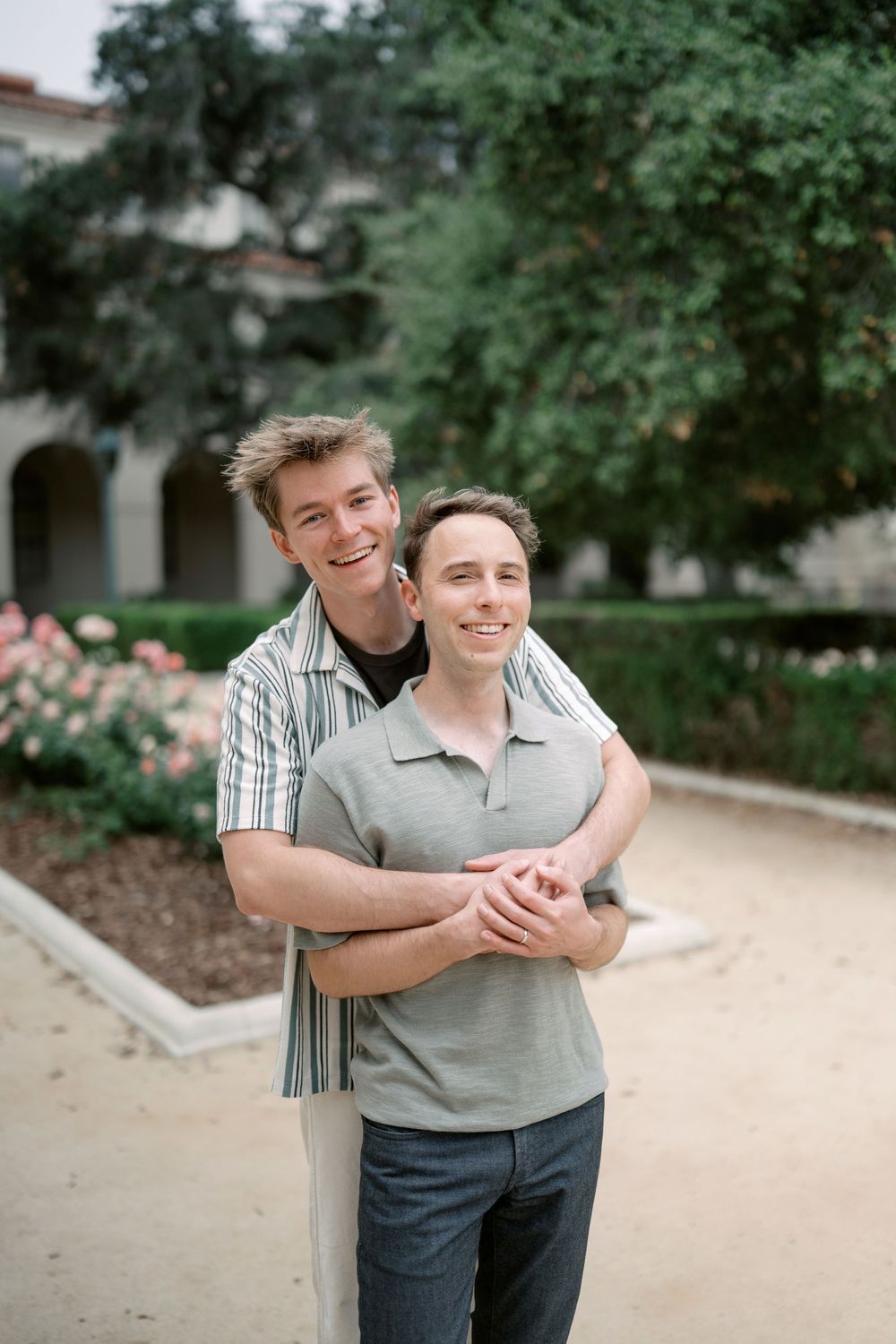 Two smiling men embrace outdoors near a garden with pink flowers and lush green trees in the background.