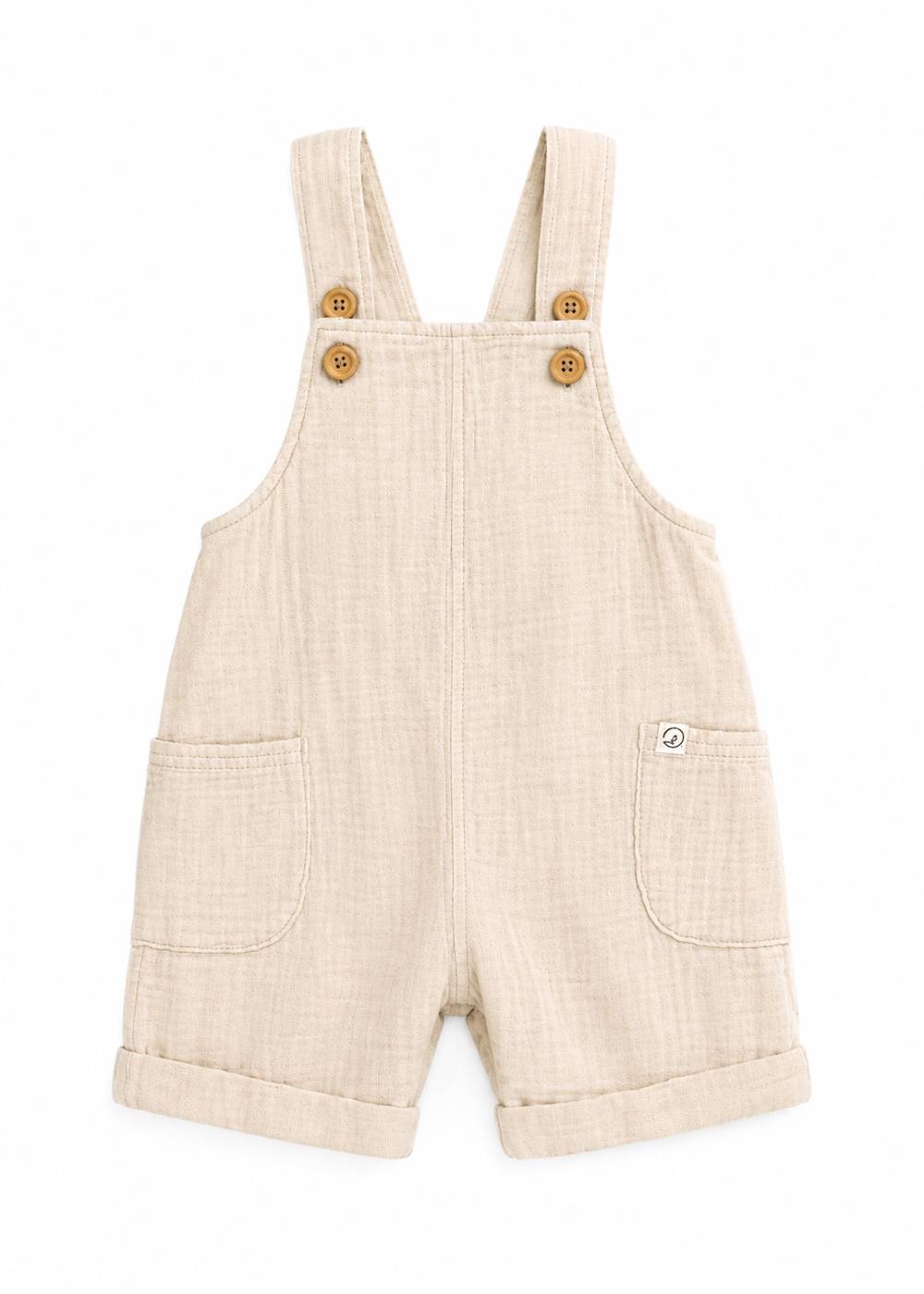 Beige cotton muslin baby dungaree shorts with gold buttons and front pockets on white background.