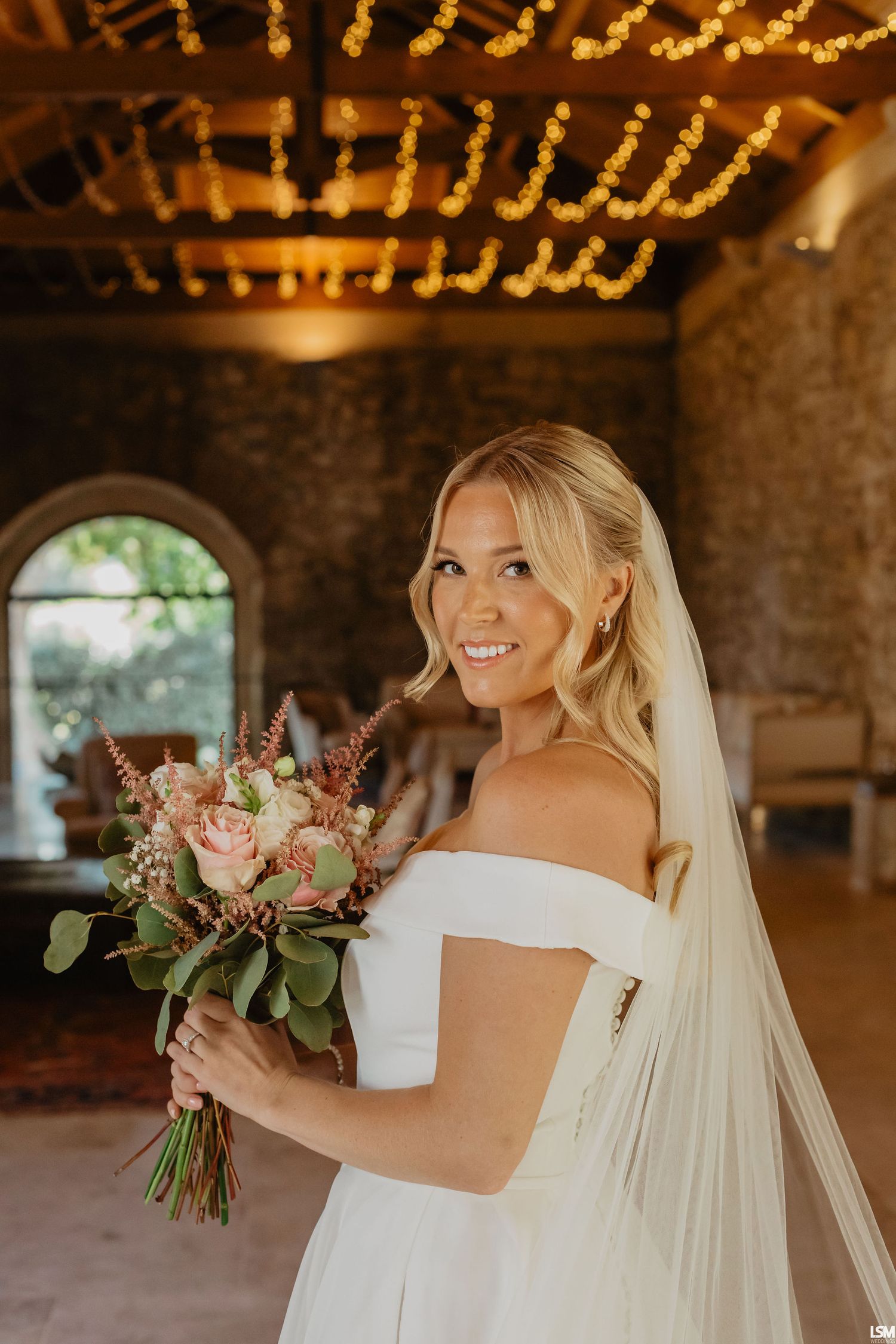 Smiling bride in off-shoulder gown holds pink bouquet, wearing veil, in rustic venue with fairy lights.