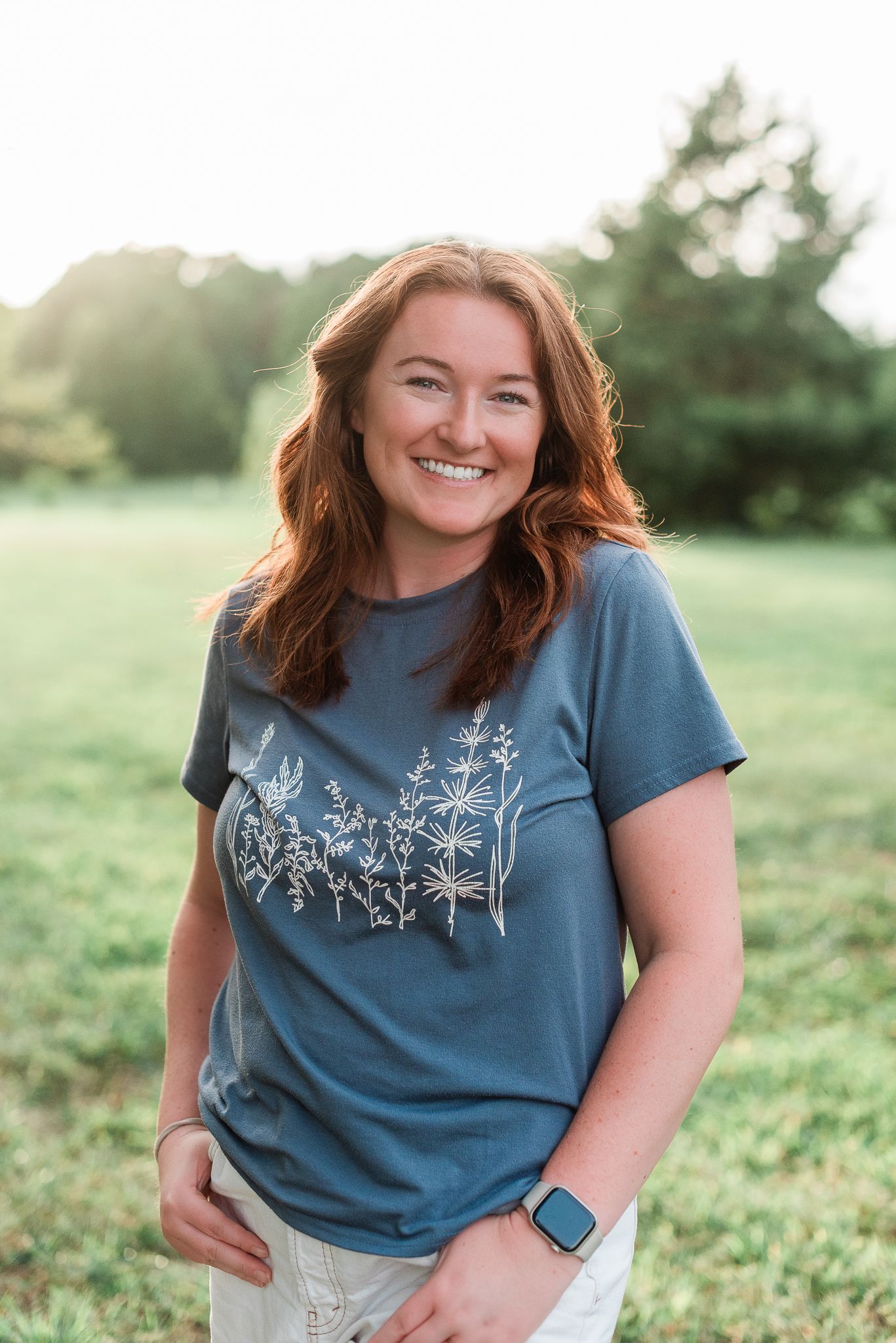 Person wearing blue t-shirt with floral design stands in grassy field during golden hour.