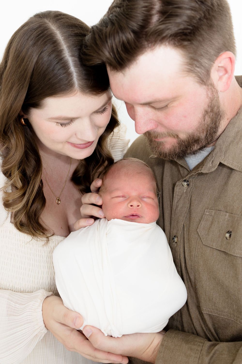 A couple tenderly gazes at their swaddled newborn baby in a bright white studio portrait.