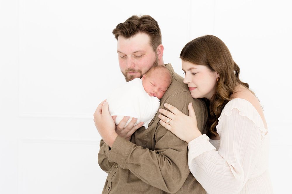 New parents tenderly hold their swaddled newborn baby together against a white studio background.