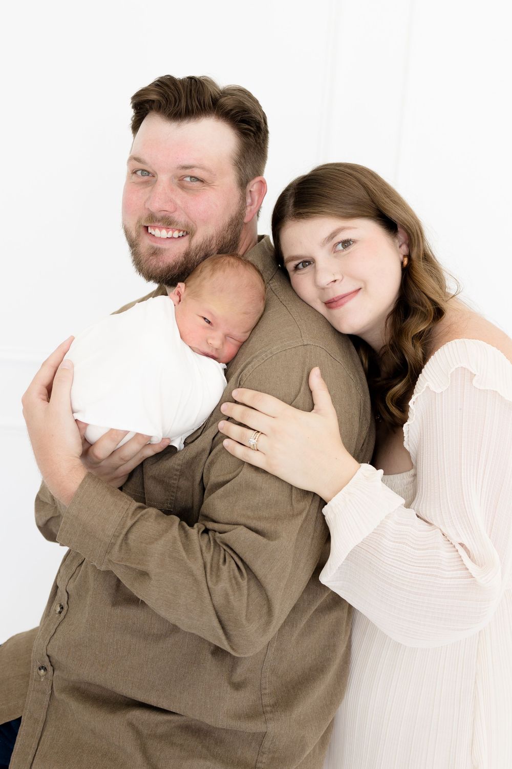Happy couple smiling and holding their newborn baby wrapped in white, posing for a family portrait against a white background.
