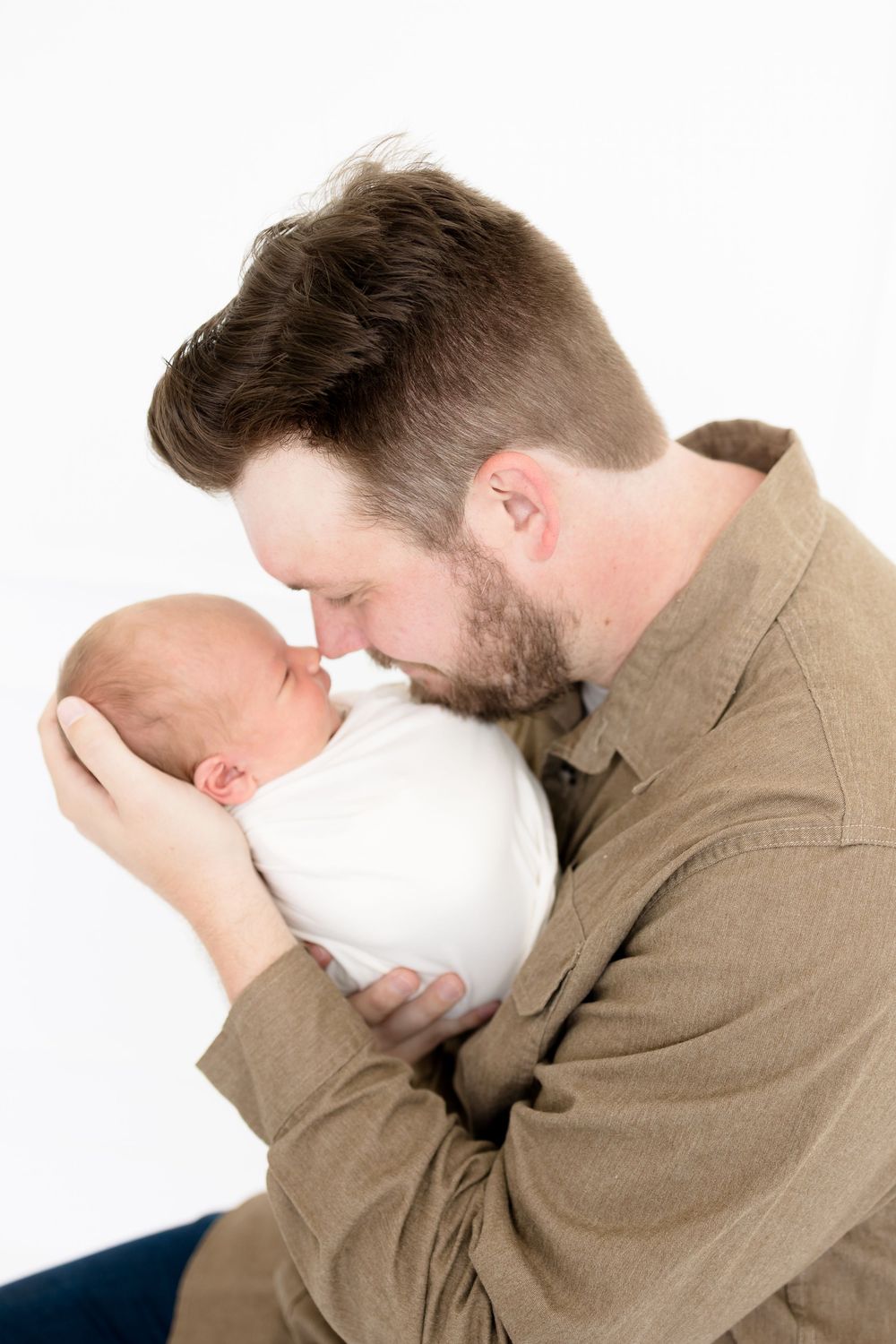 A bearded father gently touches noses with his swaddled newborn baby in a white studio photo.