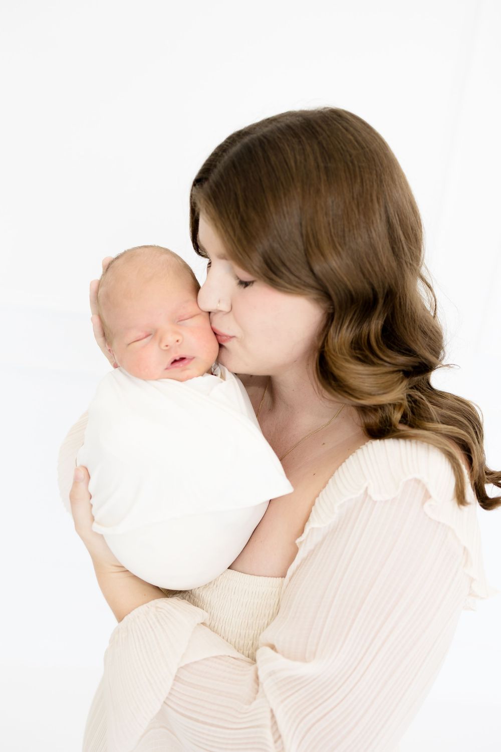 A mother gently kisses her sleeping newborn baby wrapped in white, against a soft white background.
